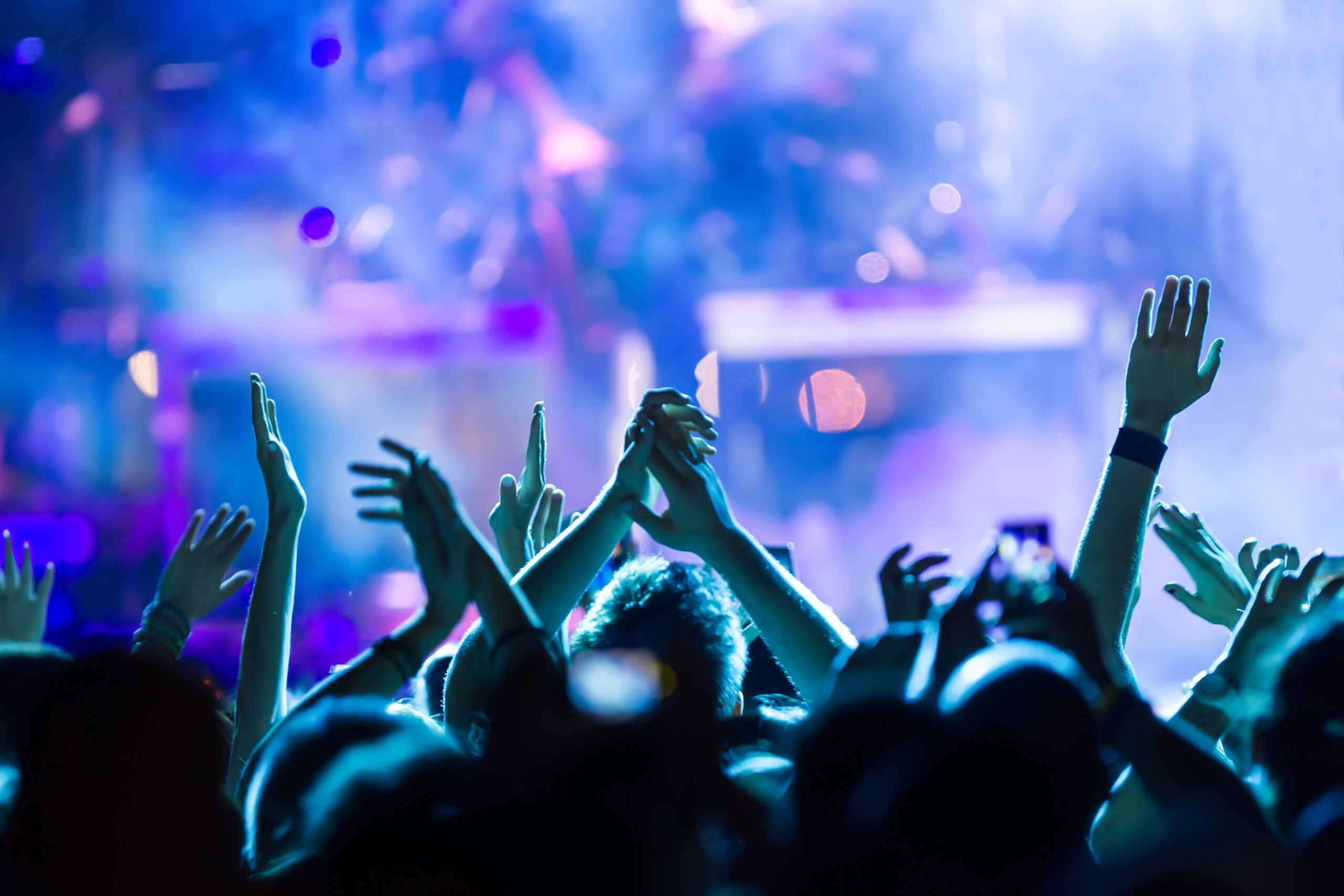 Close-up of a crowded audience with their hands raised and cheering at a live concert under vibrant blue and purple stage lights.