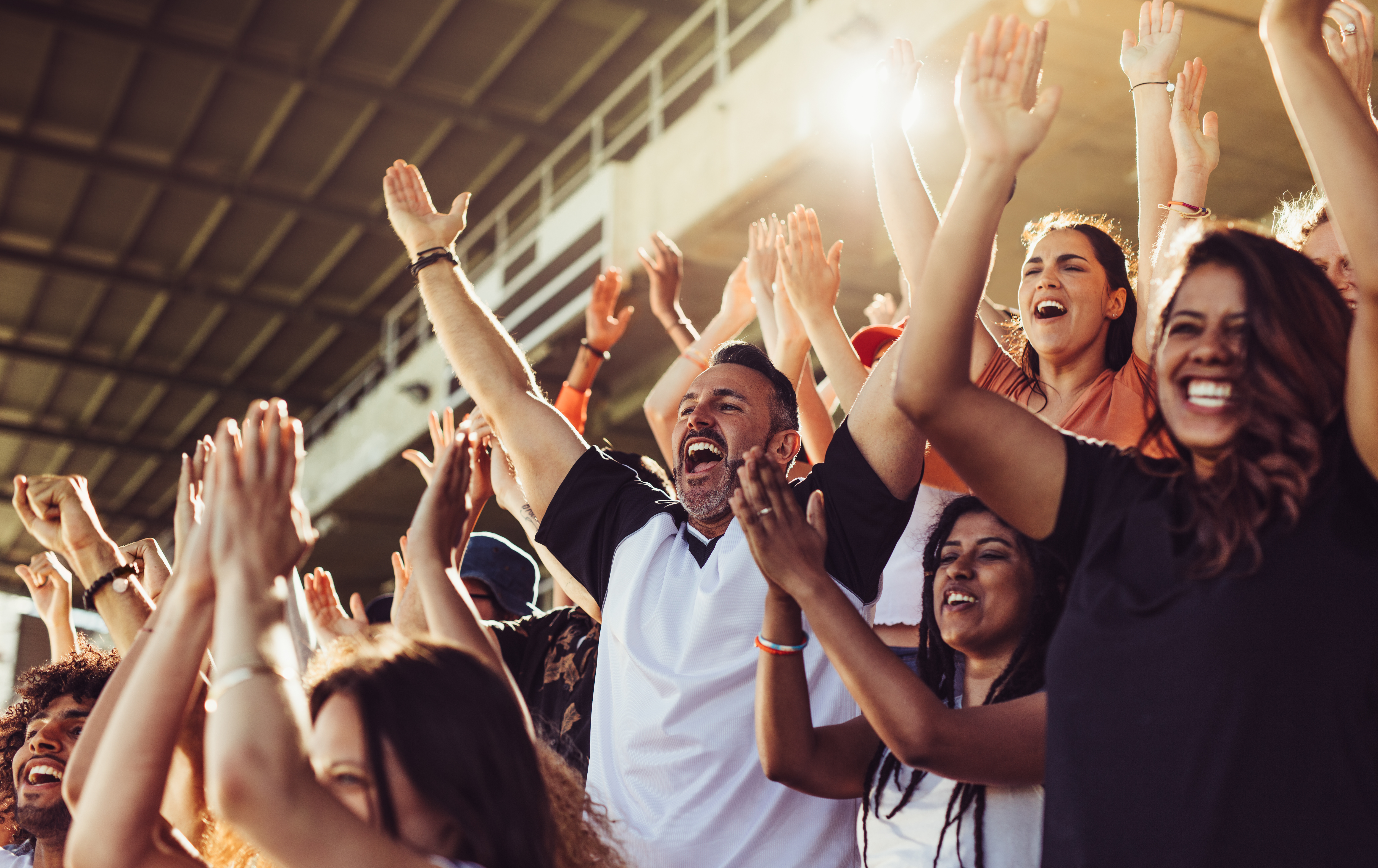 Diverse group of sports fans with hands raised, applauding and cheering loudly during a bright, action-packed stadium event.